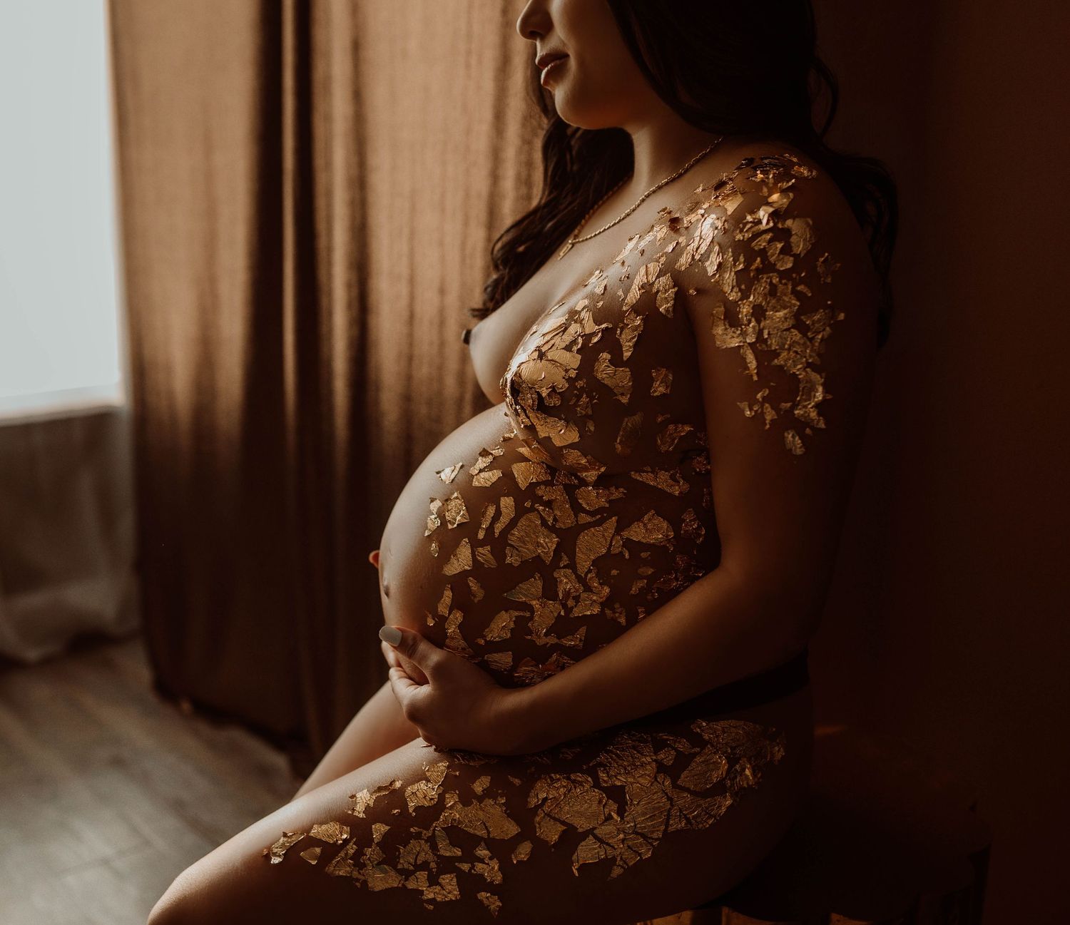 Stillwater Minnesota boudoir photoshoot, a woman cradling baby bump, seated by window with warm brown curtains.