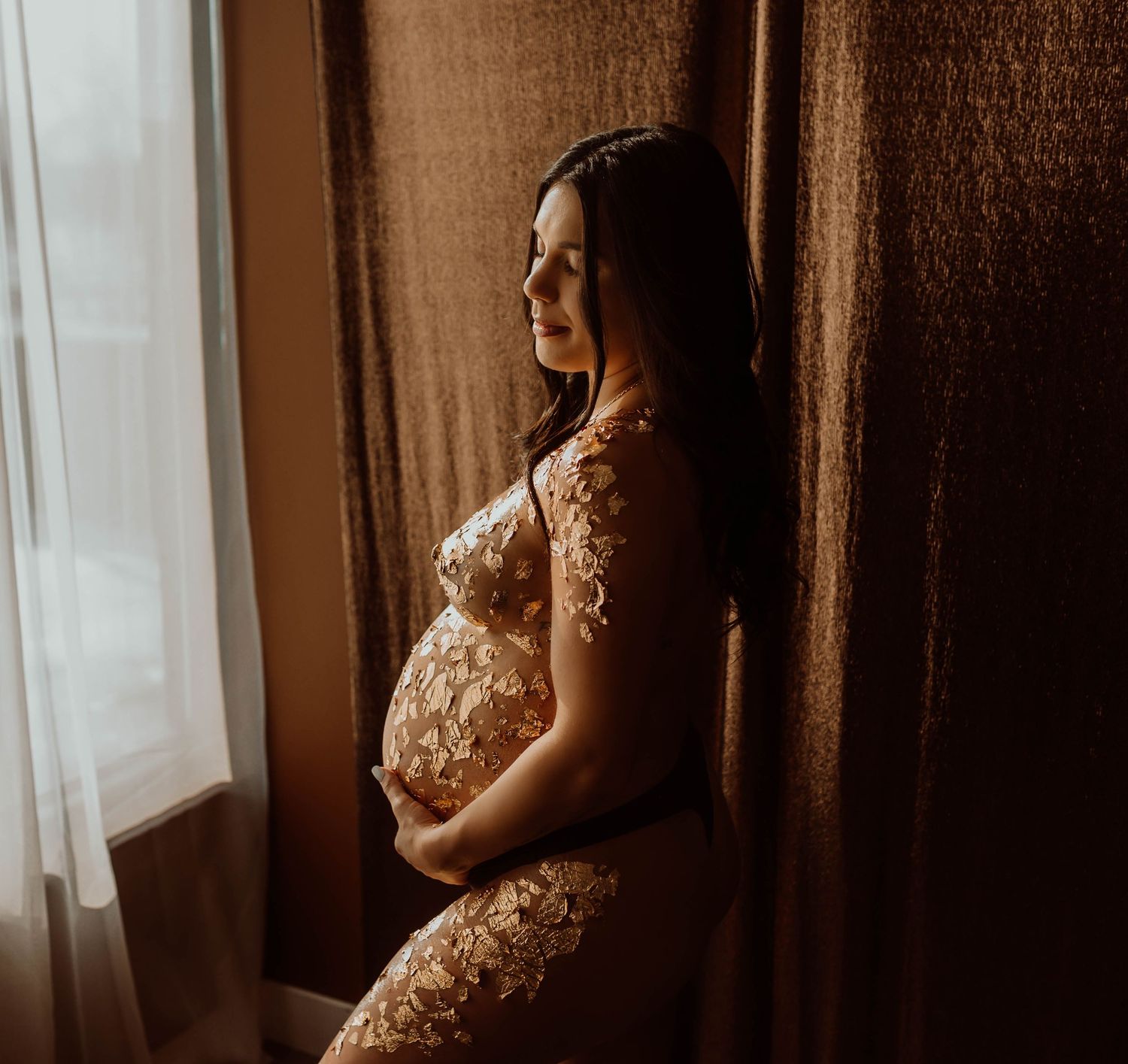 Pregnant woman in gold foil who is cradling baby bump by window with warm light and curtains - Minnesota boudoir photography.