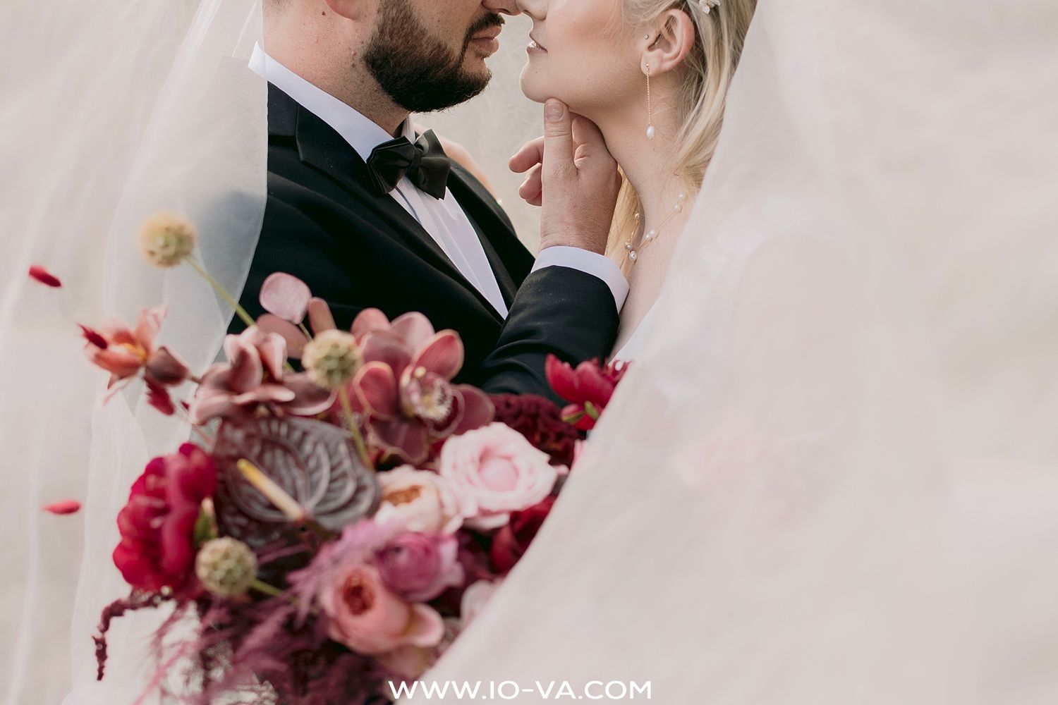 Elegant bride in off-shoulder lace wedding gown wrapped in flowing veil in soft natural light.
