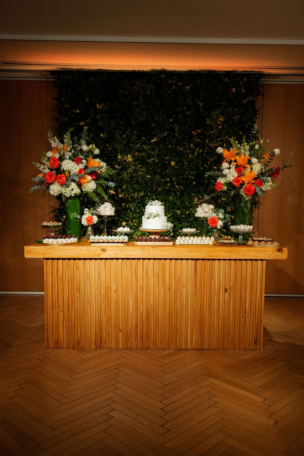 Elegant wedding dessert table with white cake, colorful floral arrangements, and green leaf backdrop.
