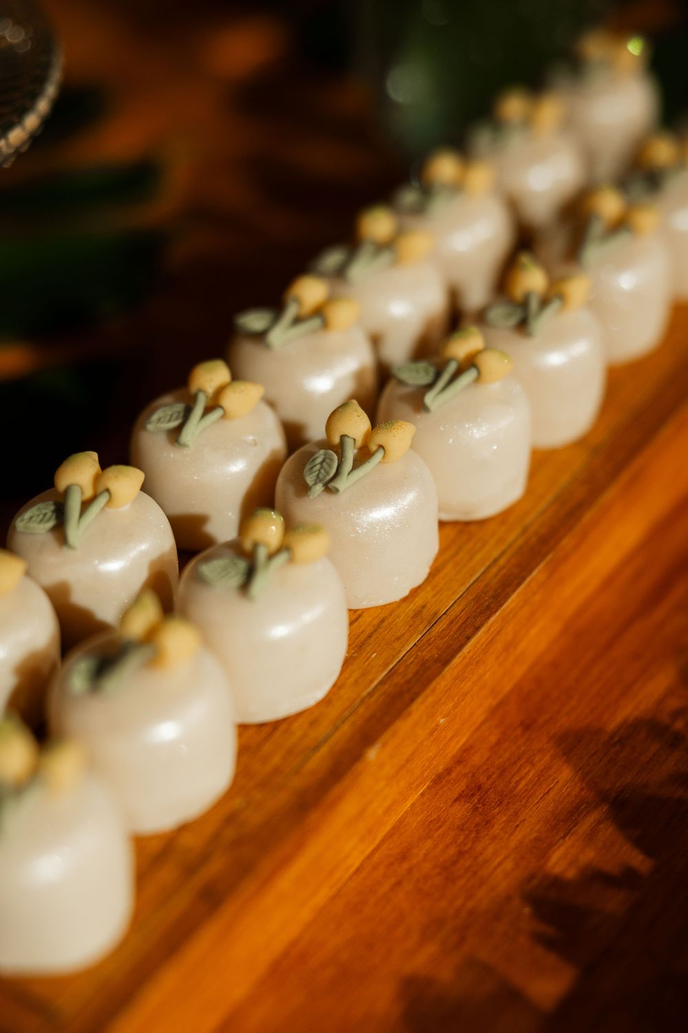 Rows of white petit four wedding favors with delicate green and yellow floral decorations on wood.
