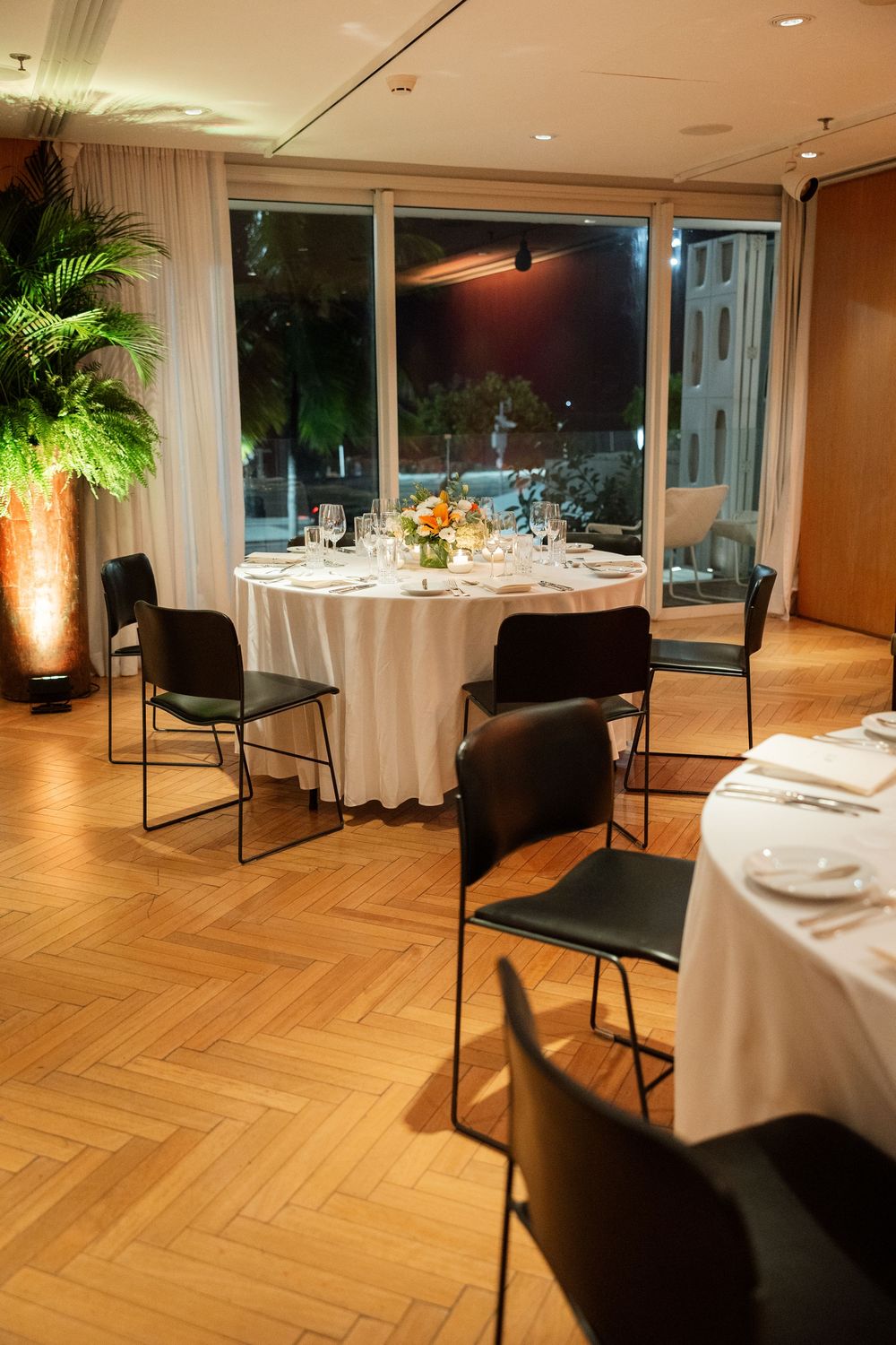 Intimate wedding reception room with round tables, black chairs, and large windows overlooking night city.