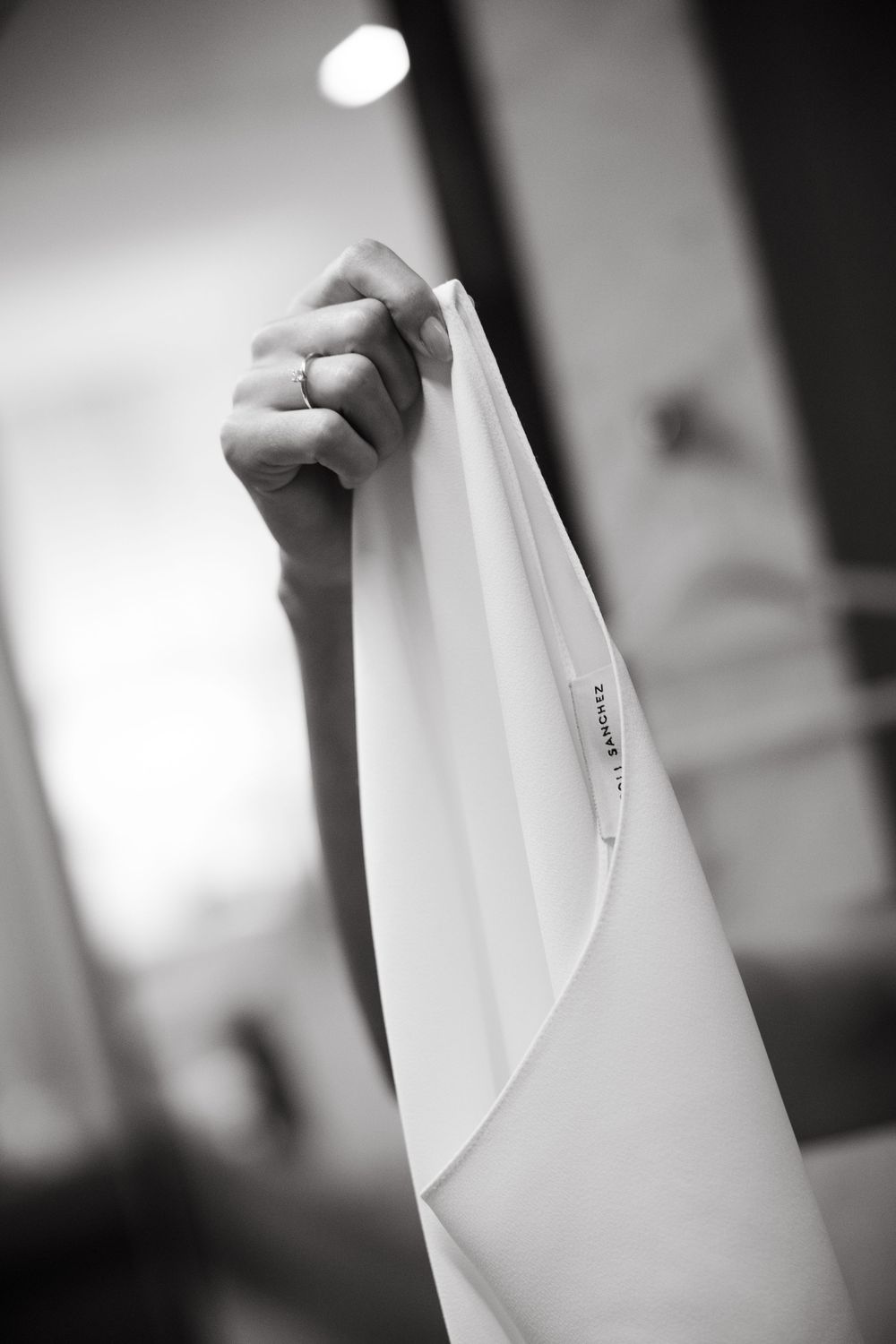 Bride holding up white wedding dress by designer label in black and white photo.