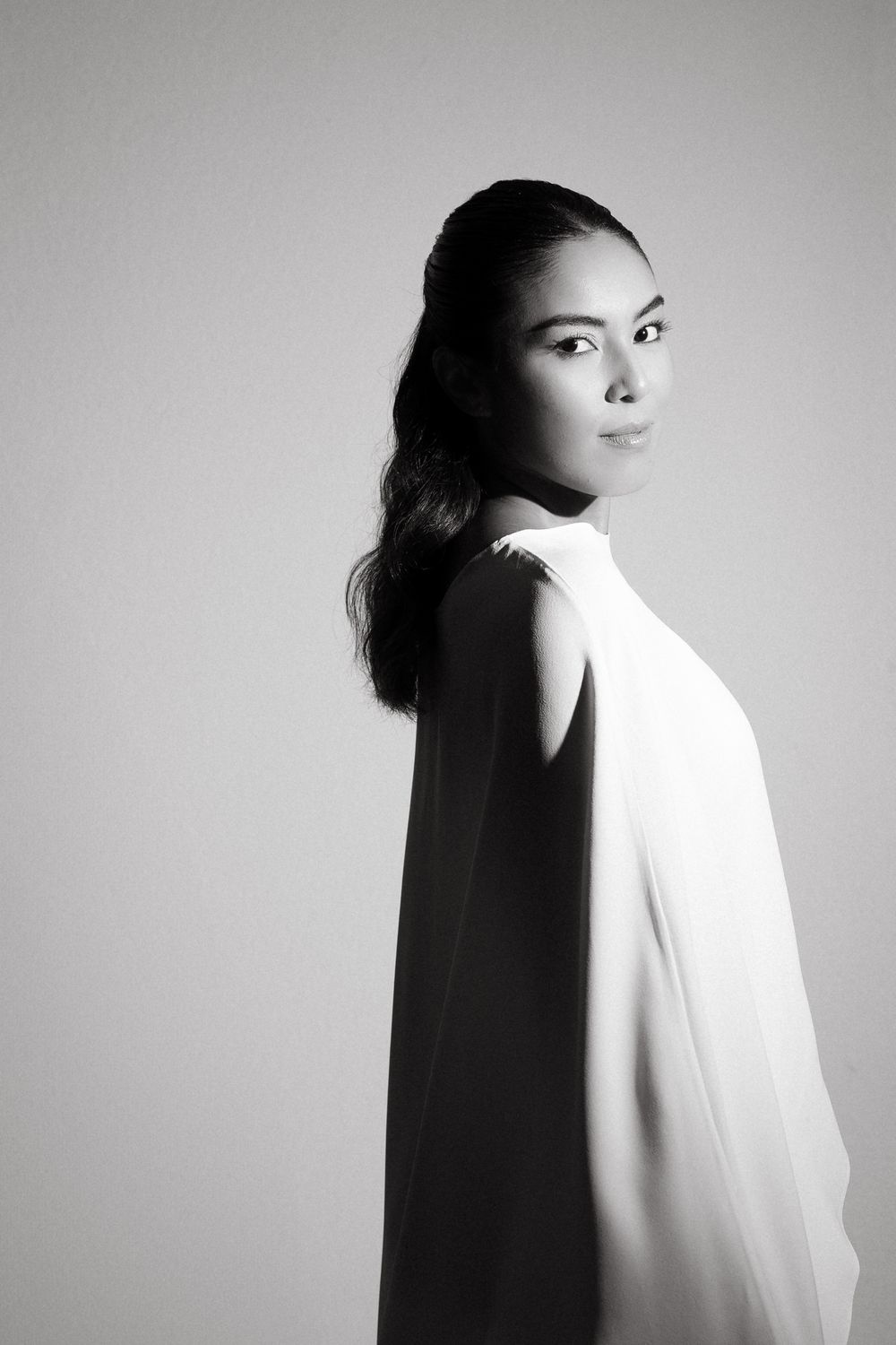 Elegant bride in white cape dress posing in dramatic black and white portrait.