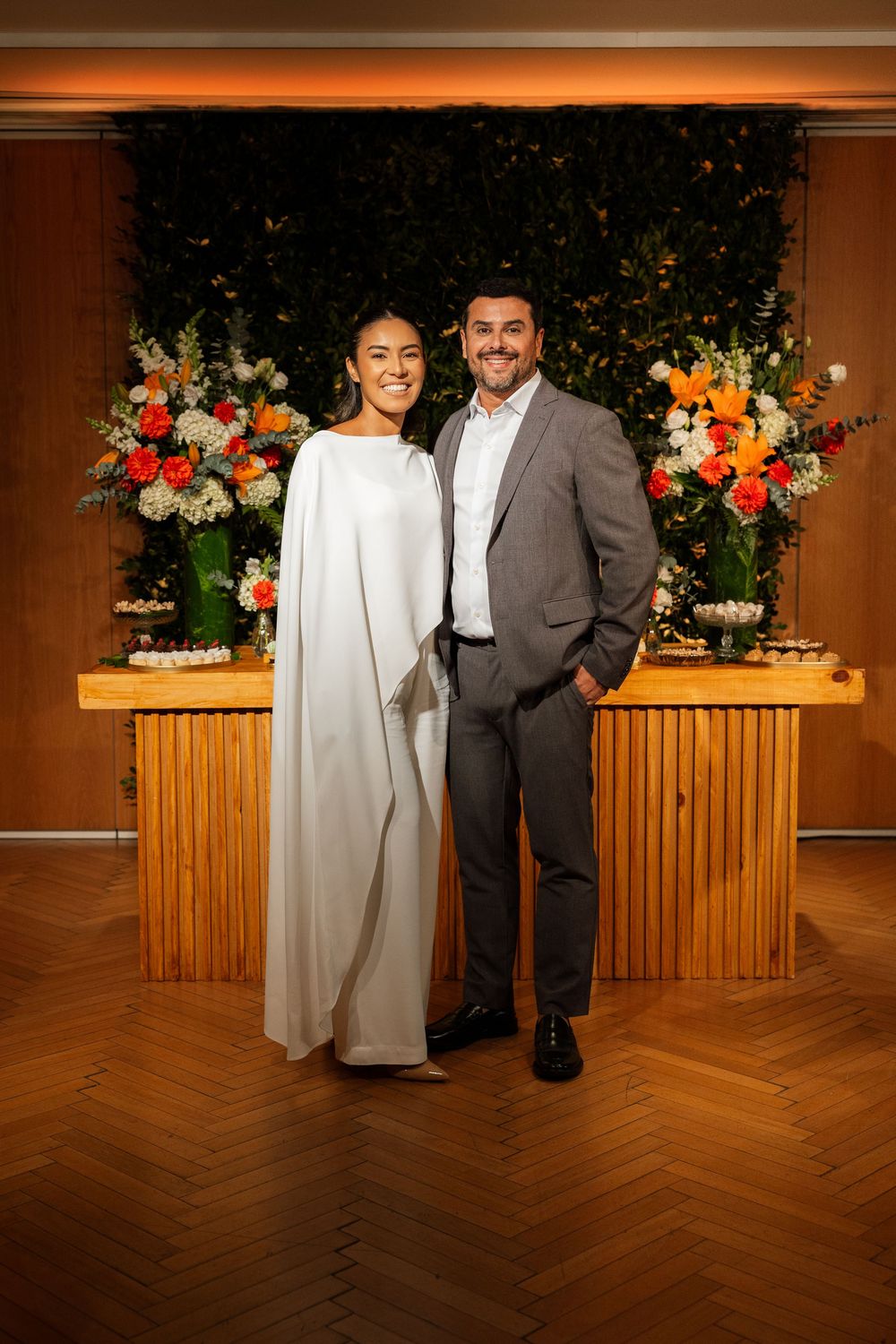 Smiling bride and groom posing before floral backdrop at wedding reception.