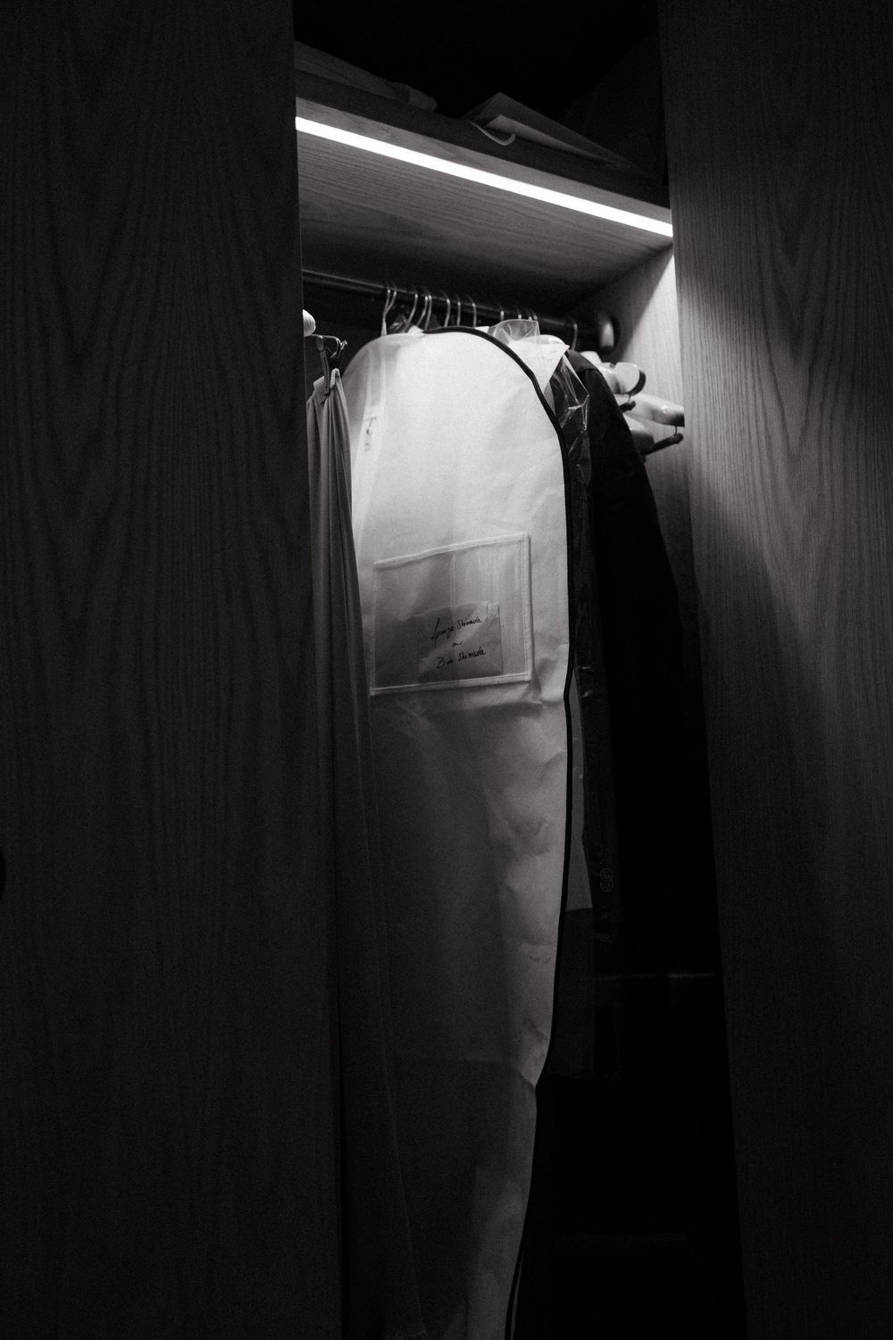 Black and white photo of a wedding dress in garment bag hanging in illuminated closet.