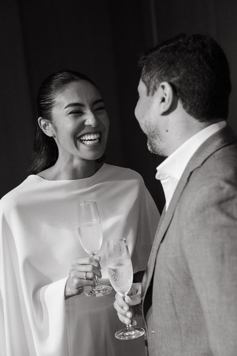 Couple laughing and toasting with champagne glasses at a wedding celebration in black and white.