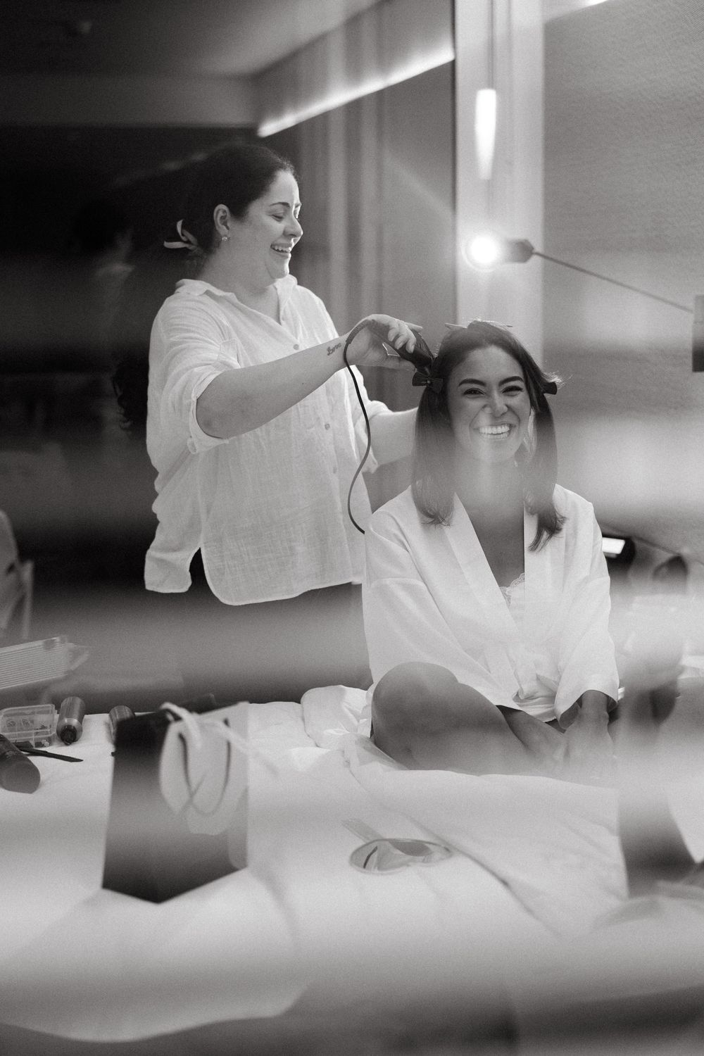 Black and white photo of bride smiling while hairstylist curls her hair before wedding ceremony.
