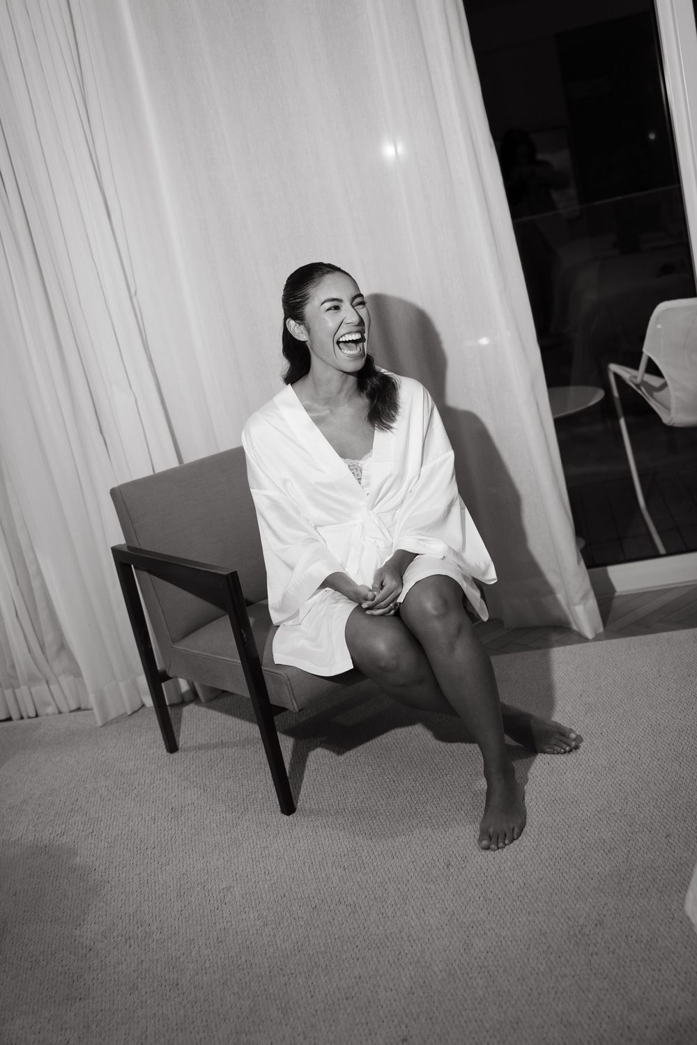 Laughing bride in white robe sitting barefoot on chair in black and white photo.