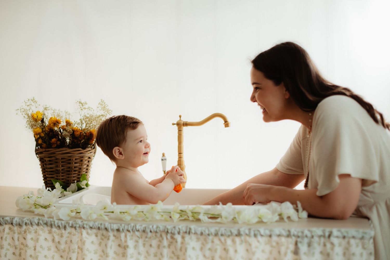 A mother smiles at her happy baby sitting in a flower-decorated bathtub with a golden faucet.