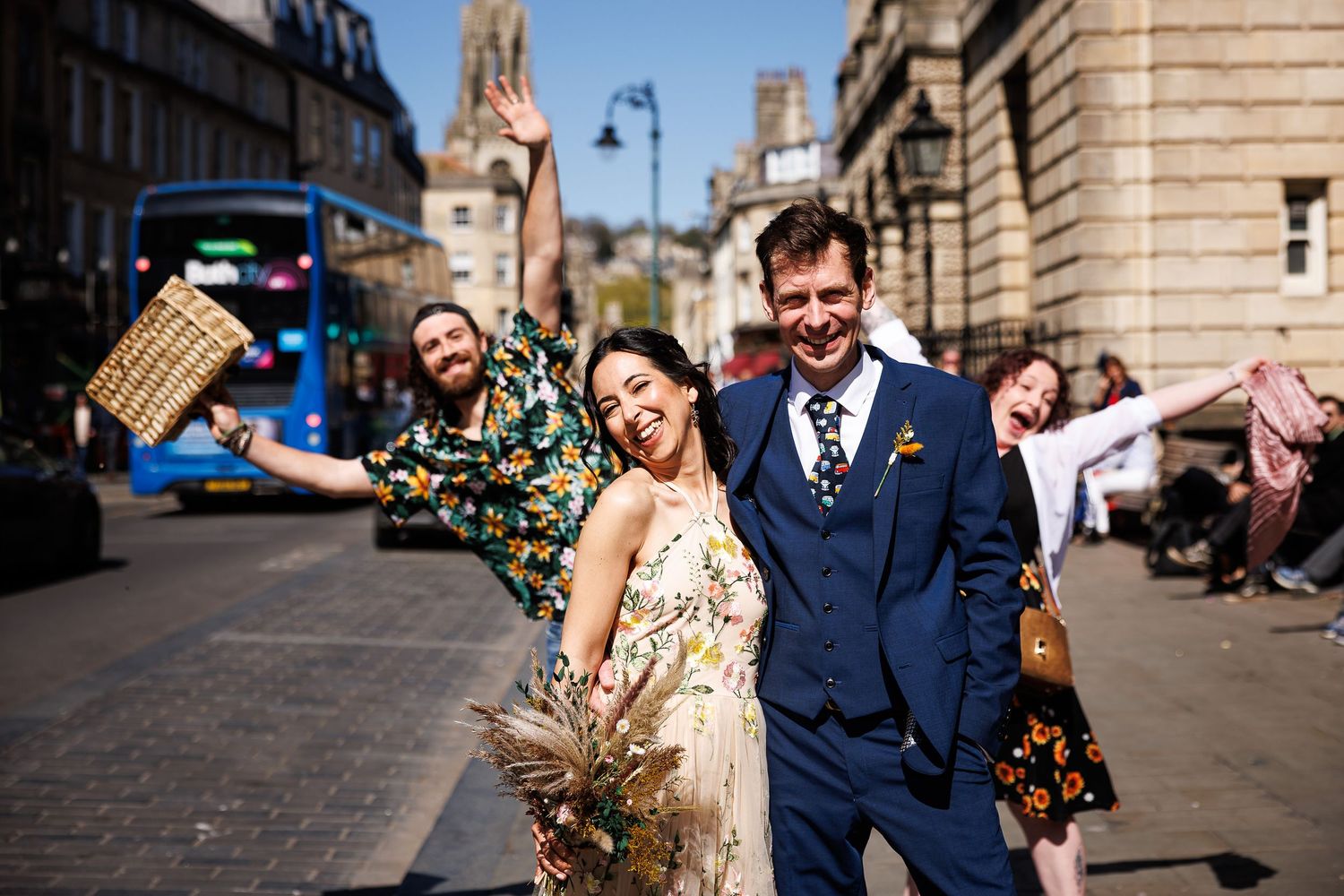 Joyful couple celebrating their wedding on a sunny Edinburgh street with friends cheering behind.