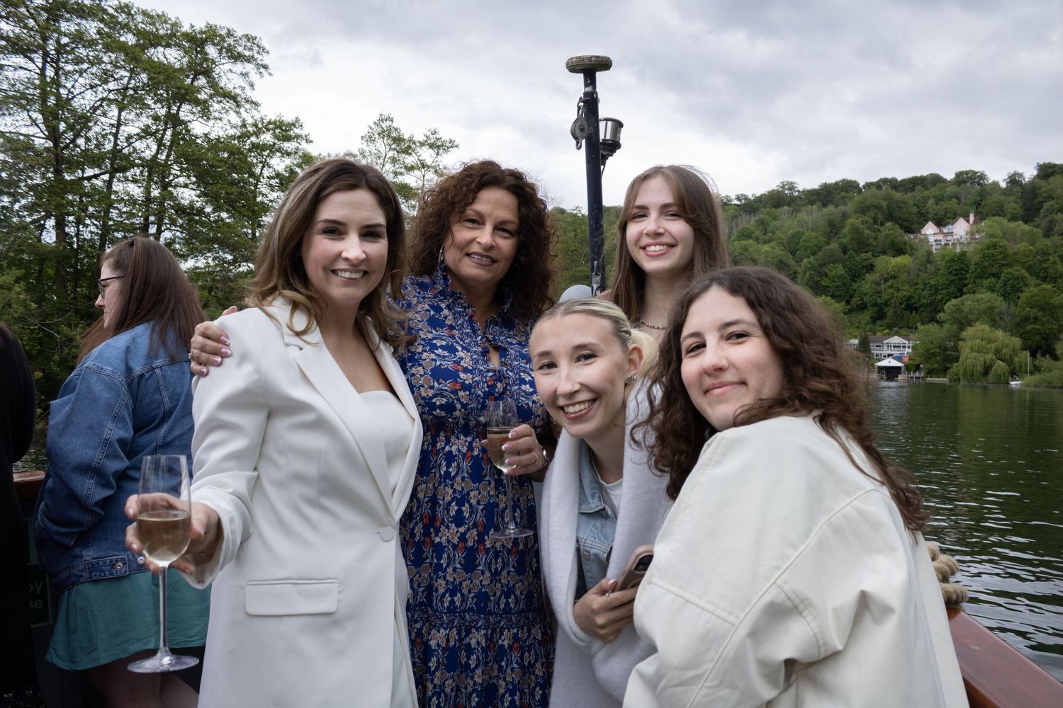 Group of five women smiling together outdoors near a lake at an event.