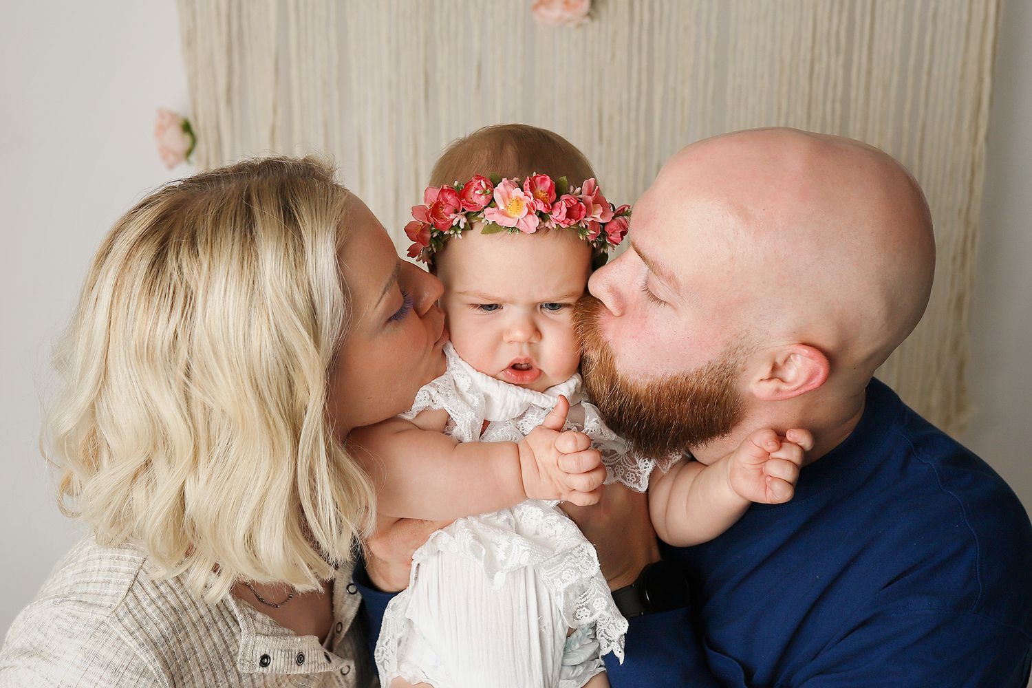 Parents kissing their baby girl wearing a floral headband in a soft, bright studio family portrait.