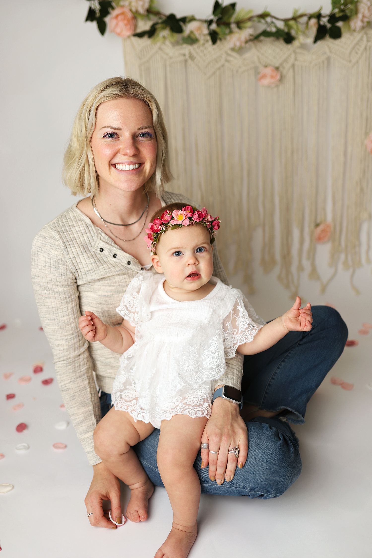 A smiling blonde mother sits on a white floor holding her baby girl in a white lace dress and floral crown.