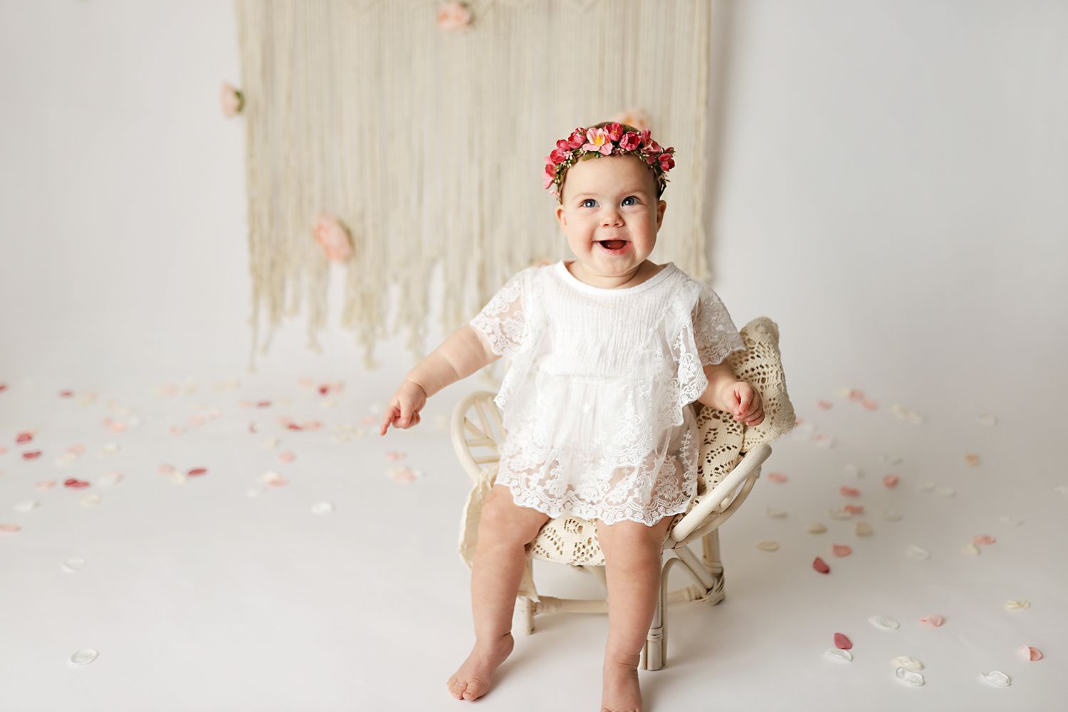 Baby girl in white lace romper and floral crown sits on chair surrounded by rose petals in bright studio. Utah County cake smash photography