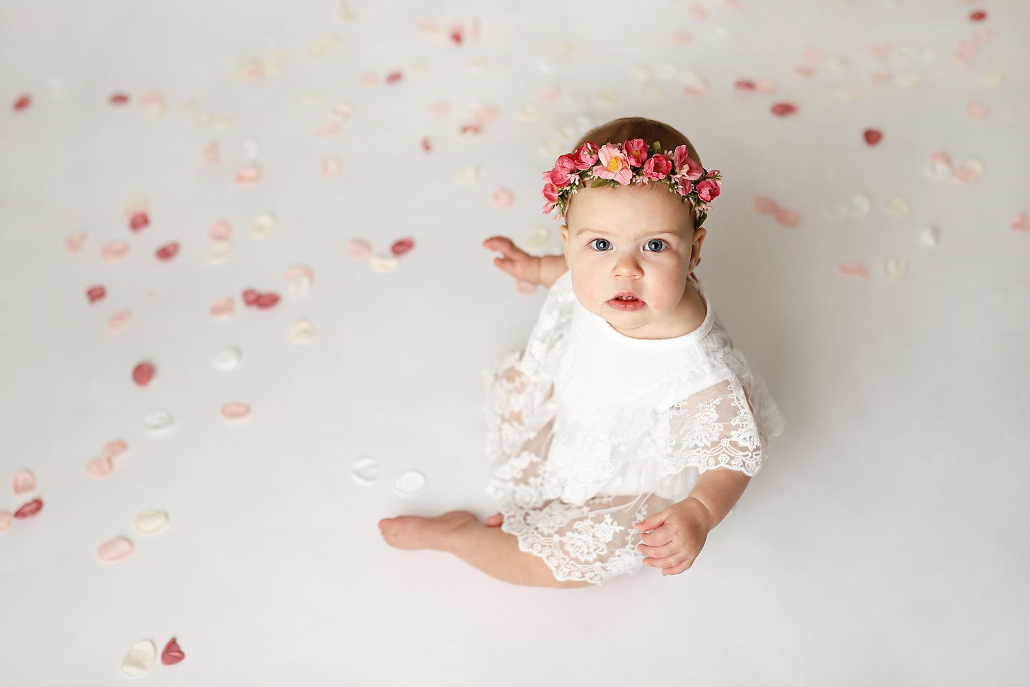 Baby girl in white lace dress and floral crown sits among rose petals on white background.
