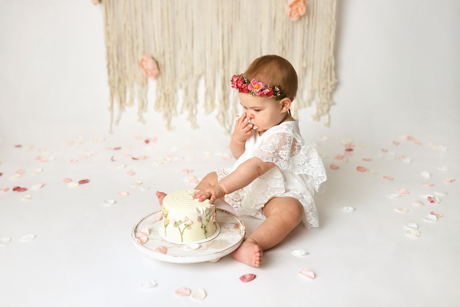 Baby girl in white lace romper smashing first birthday cake on white backdrop with floral confetti. Utah cake smash photographer