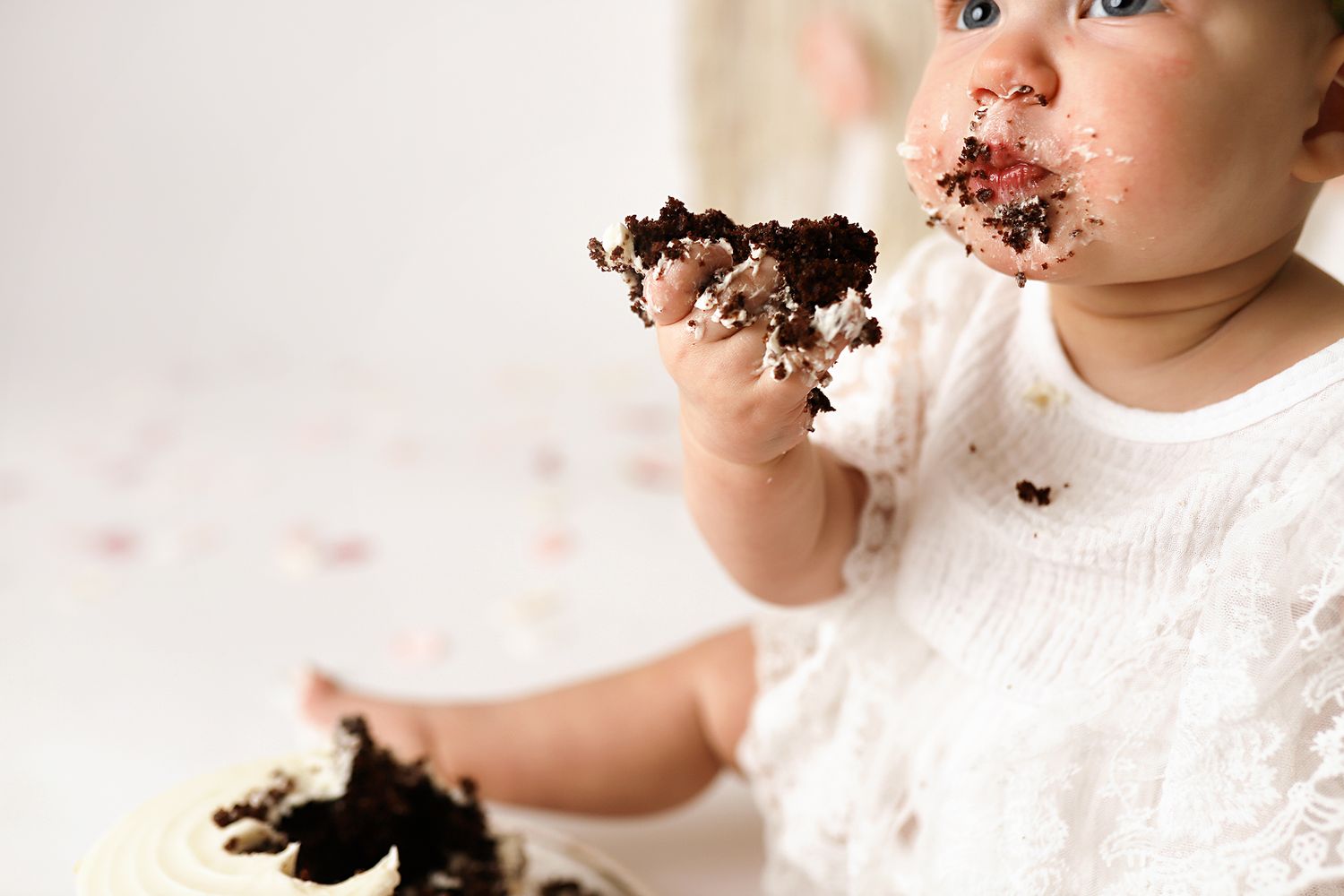 Baby girl in white dress smashing chocolate birthday cake, face and hands covered in frosting during first birthday celebration.