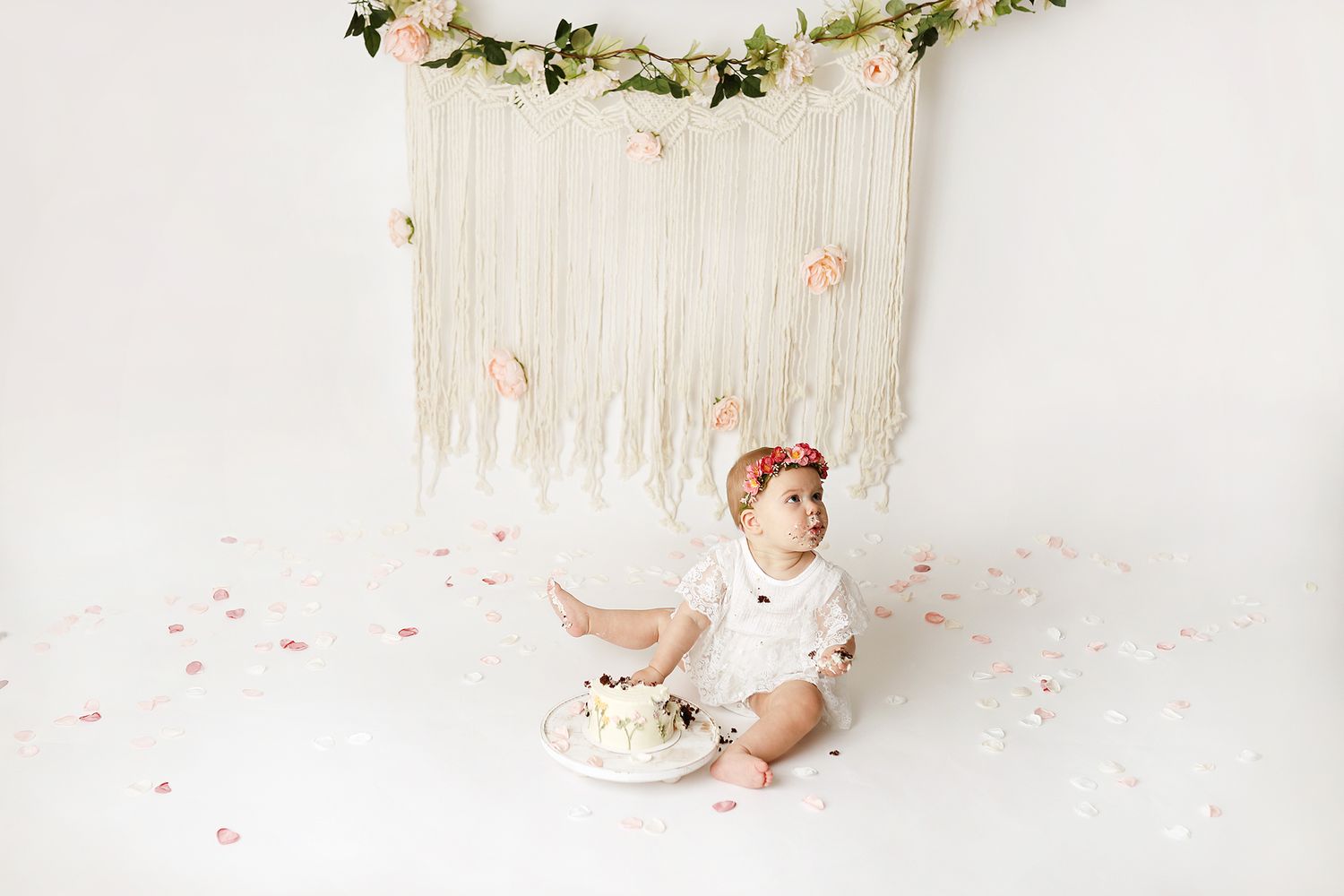 Baby girl in white outfit sits on floor with smash cake, floral crown, confetti, and boho macrame backdrop. Orem cake smash photographer