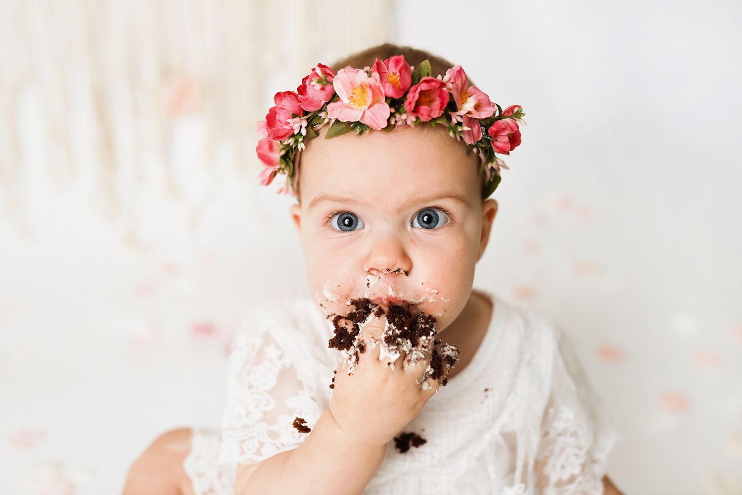 Baby girl in floral crown eating birthday cake smash, wearing white lace dress with blue eyes. Utah County cake smash photographer