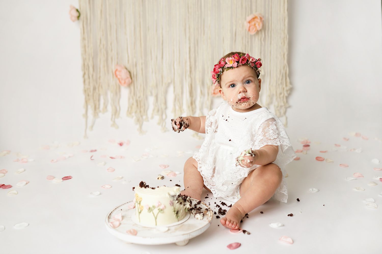 Baby girl in white lace dress and floral crown smashing her first birthday cake on white backdrop. Orem cake smash photographer.