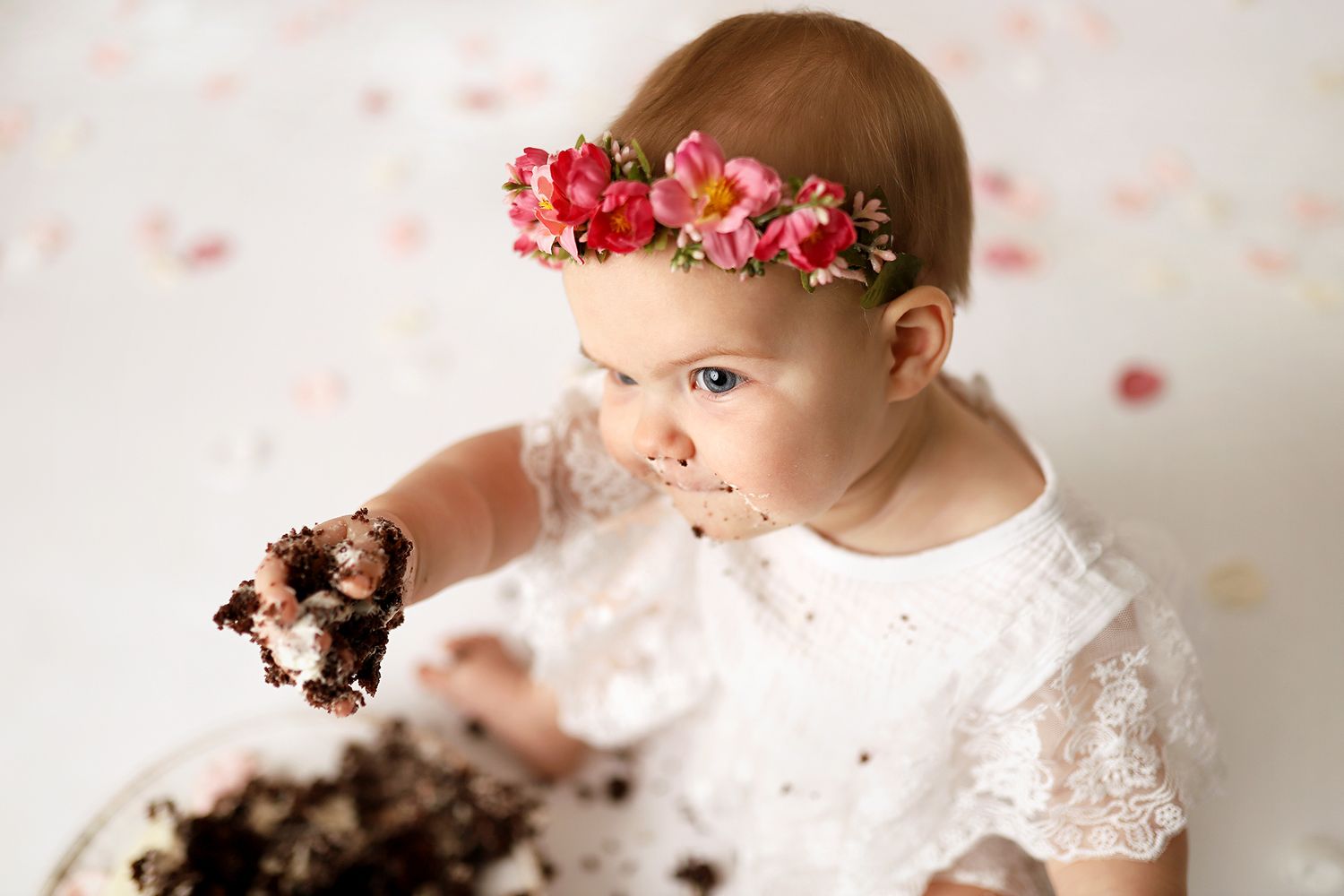 Baby girl in white lace dress and floral crown smashing chocolate birthday cake during first birthday photoshoot.