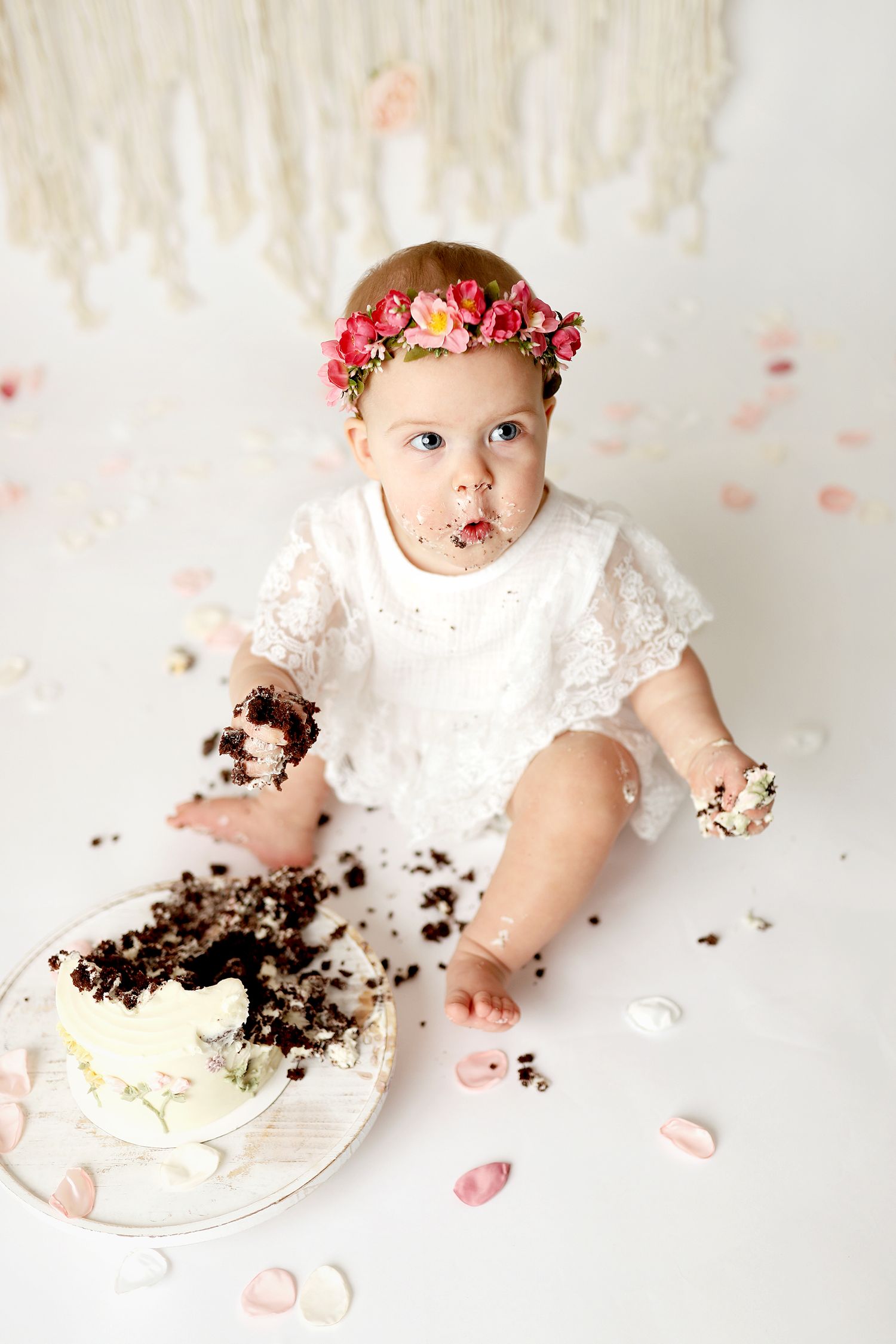 Baby girl in white lace dress and floral crown smashing first birthday cake on white background.