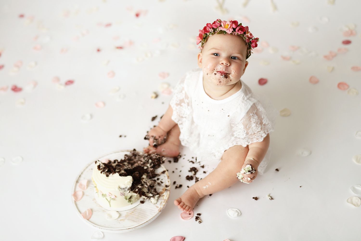 Baby girl in white lace romper and floral crown smashing first birthday cake on white background. Utah County cake smash photographer