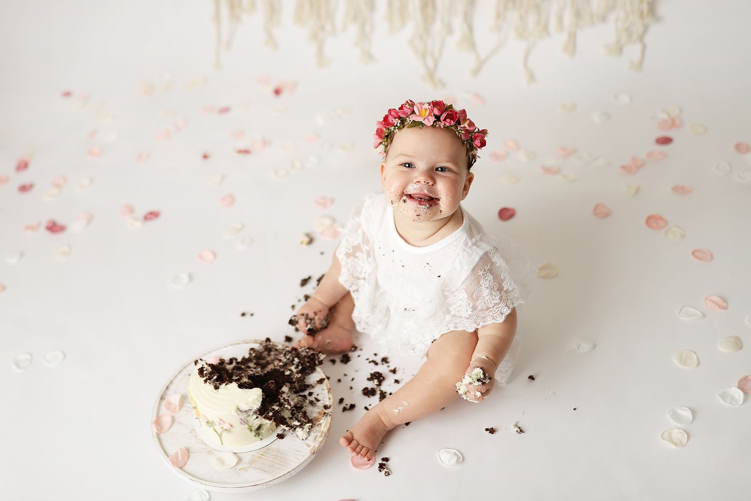 Baby girl in white lace outfit and floral crown smashing her first birthday cake on a white background.