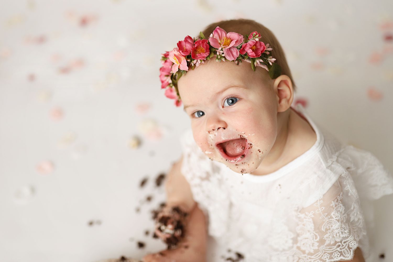 Smiling baby girl with floral crown and cake-covered face during first birthday cake smash photo.