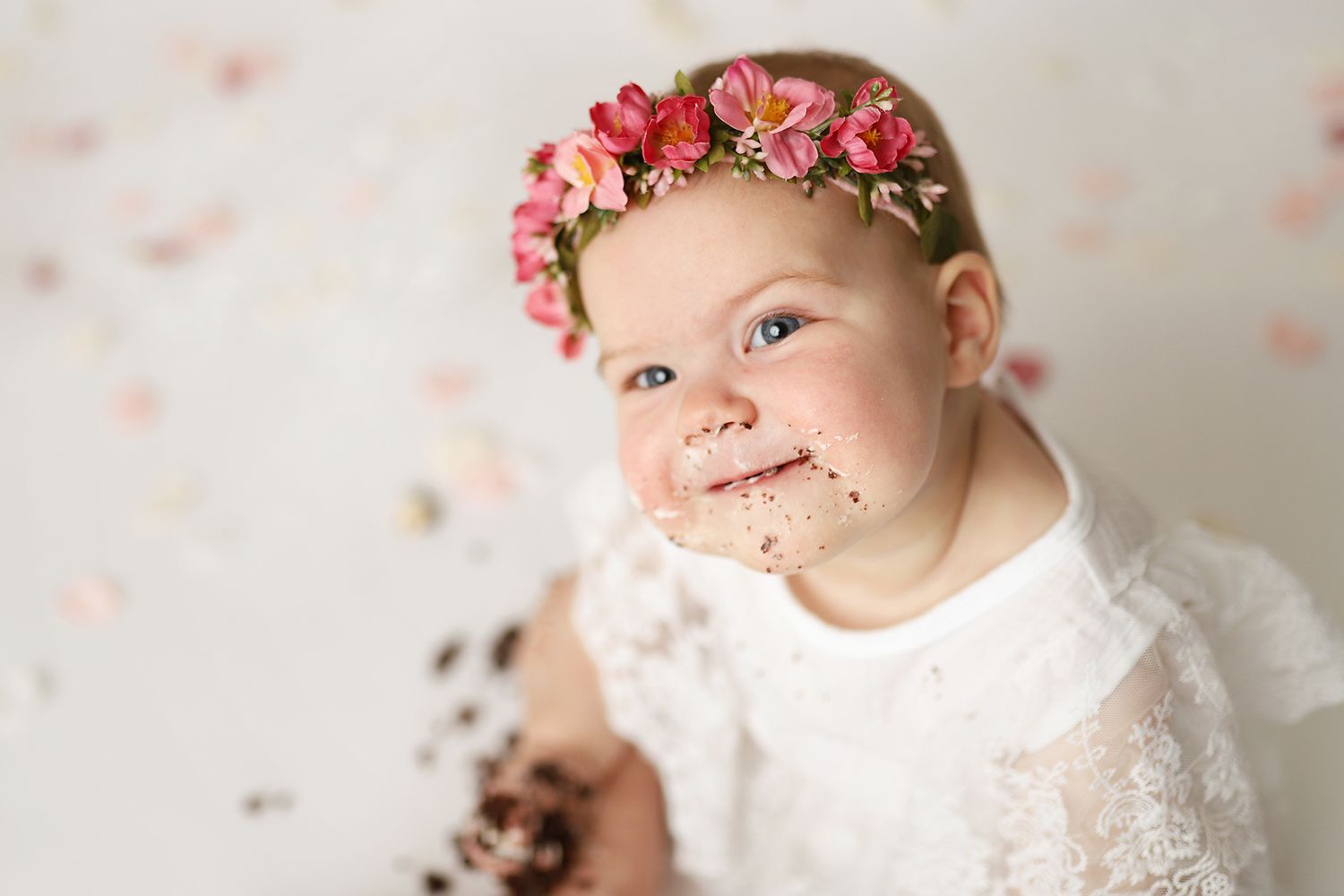 Baby girl in white lace dress and pink floral crown smiles with chocolate cake on her face during first birthday smash.