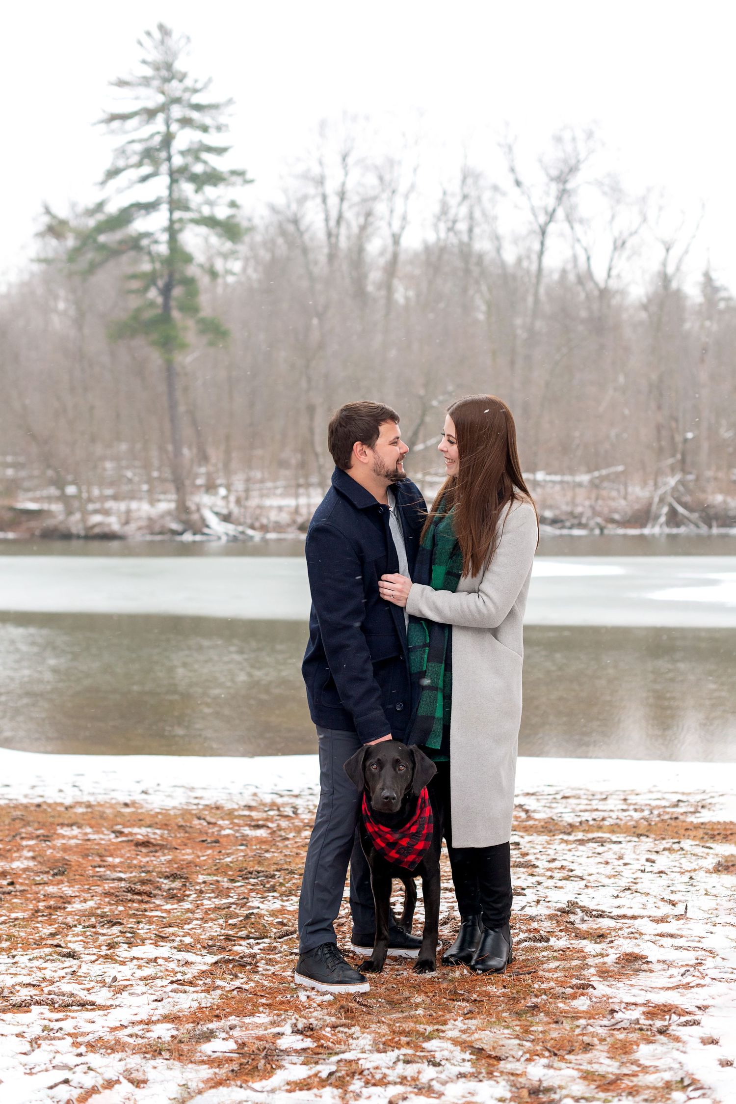 Couple gazing at each other with black Labrador wearing plaid bandana, standing by snowy frozen lake in winter.