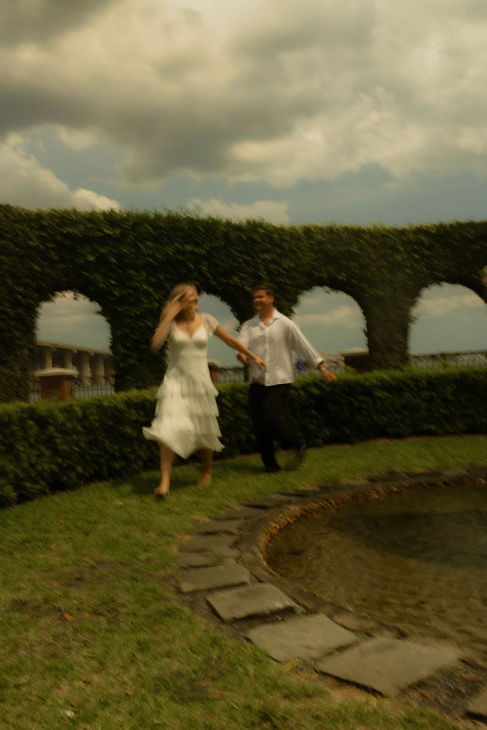Blurry photo of a couple holding hands and running through a lush garden with arched hedges under a cloudy sky.