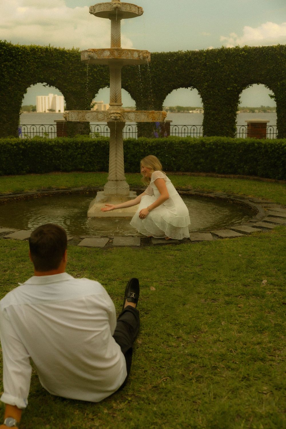 Couple in white attire near a stone garden fountain surrounded by ivy-covered arches and lush green hedges.