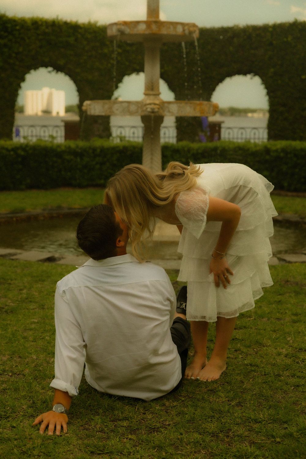 Couple in white attire sharing a romantic moment by an ornate garden fountain on green grass.