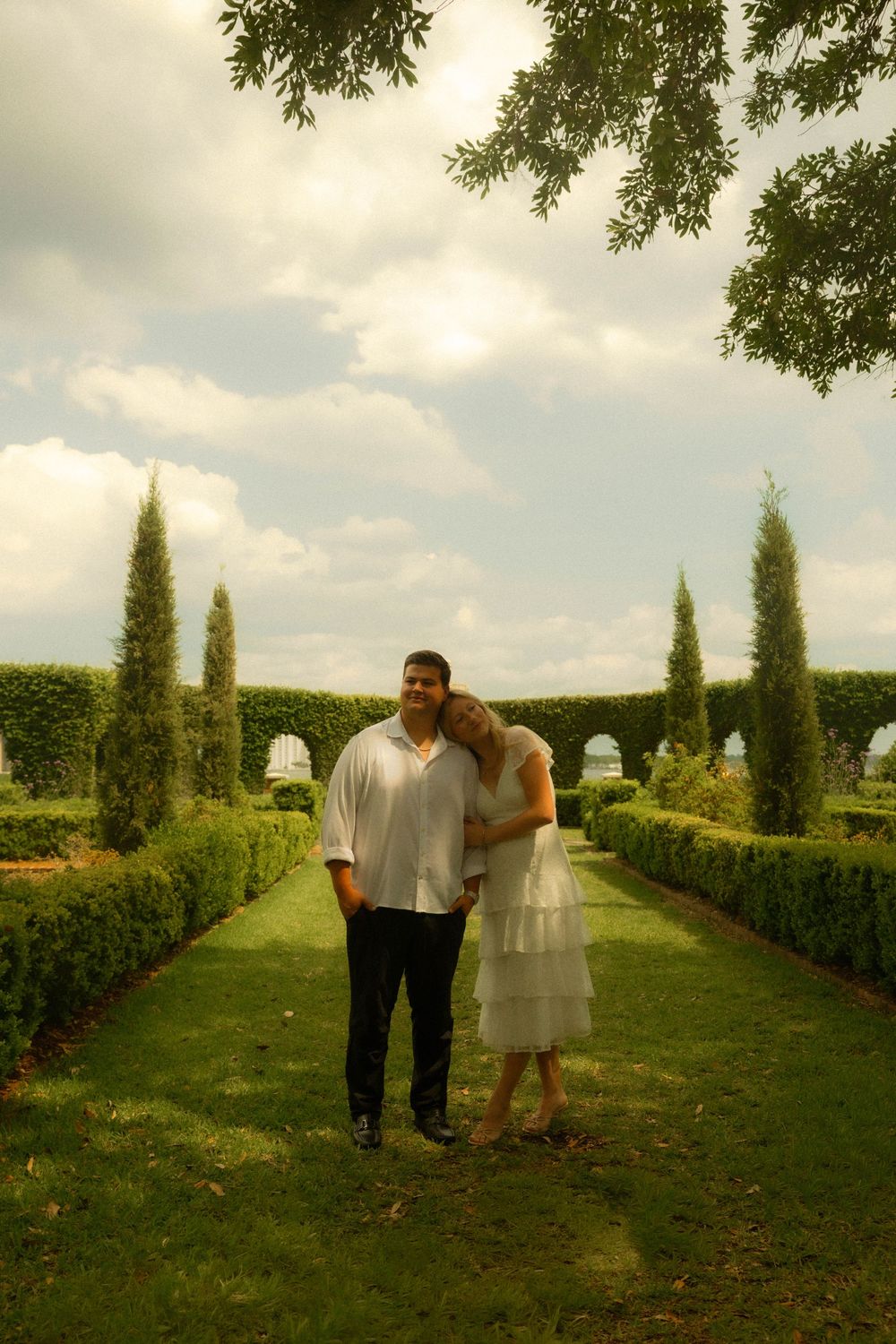 Couple in white attire standing together in a formal garden with tall cypress trees and manicured hedges.