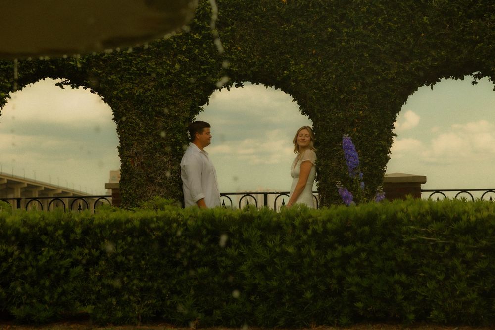 Couple standing in a lush green garden with arched hedges and purple flowers under a cloudy sky.