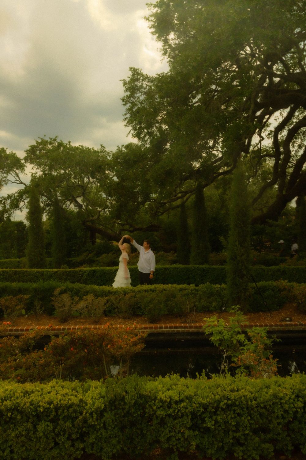A bride and groom dance among lush green hedges and towering oak trees in a romantic garden setting.