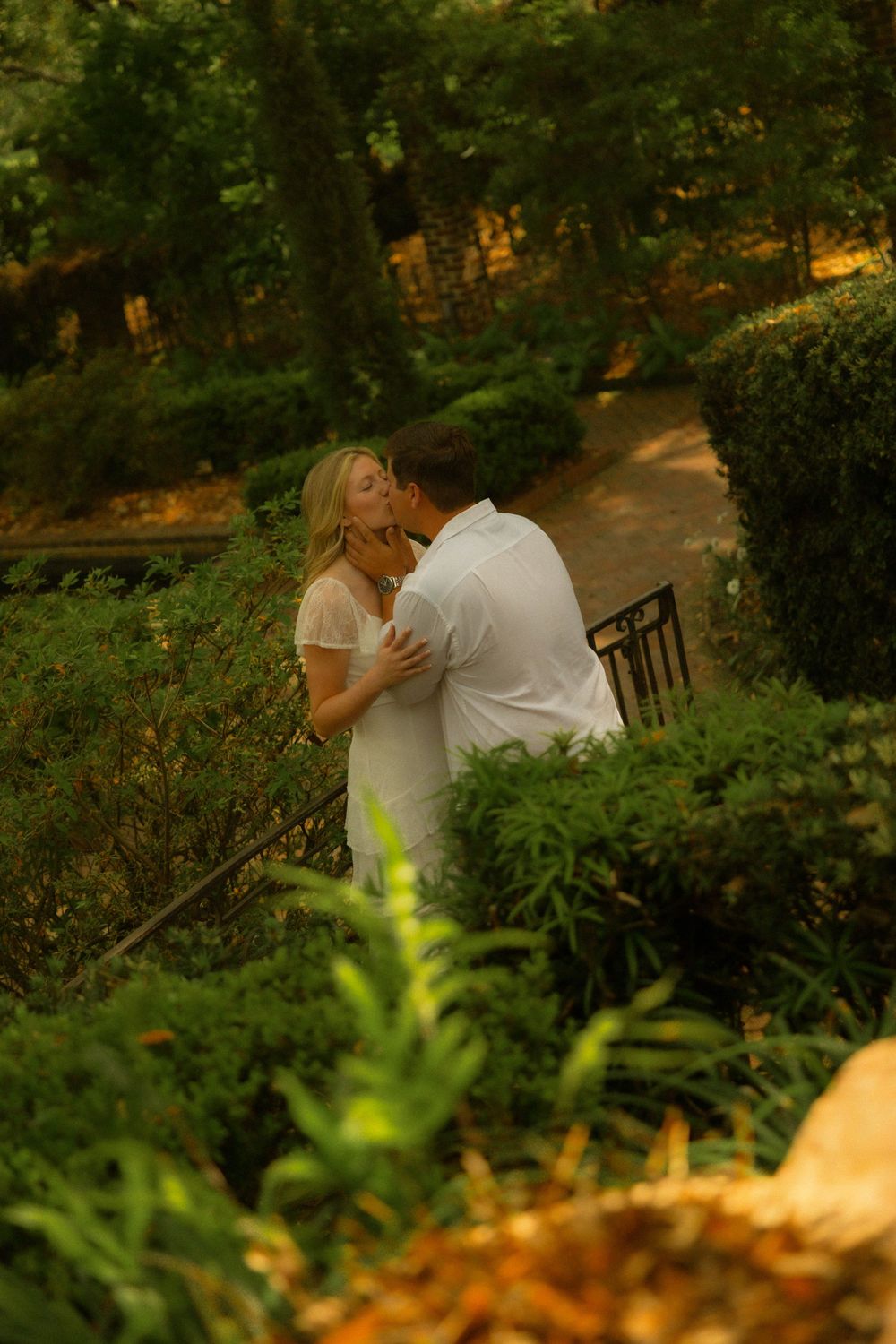 Couple sharing a romantic kiss in a lush green garden, woman in white dress embraced by man in white shirt.