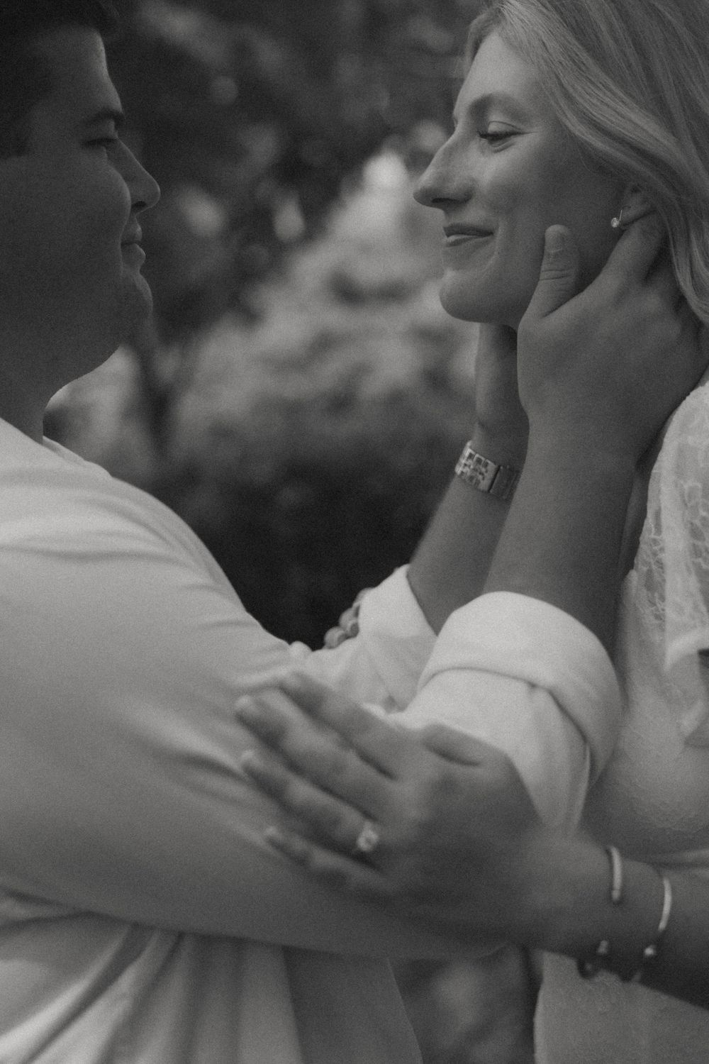 Black and white romantic photo of a couple embracing outdoors, man tenderly touching woman's face near garden.