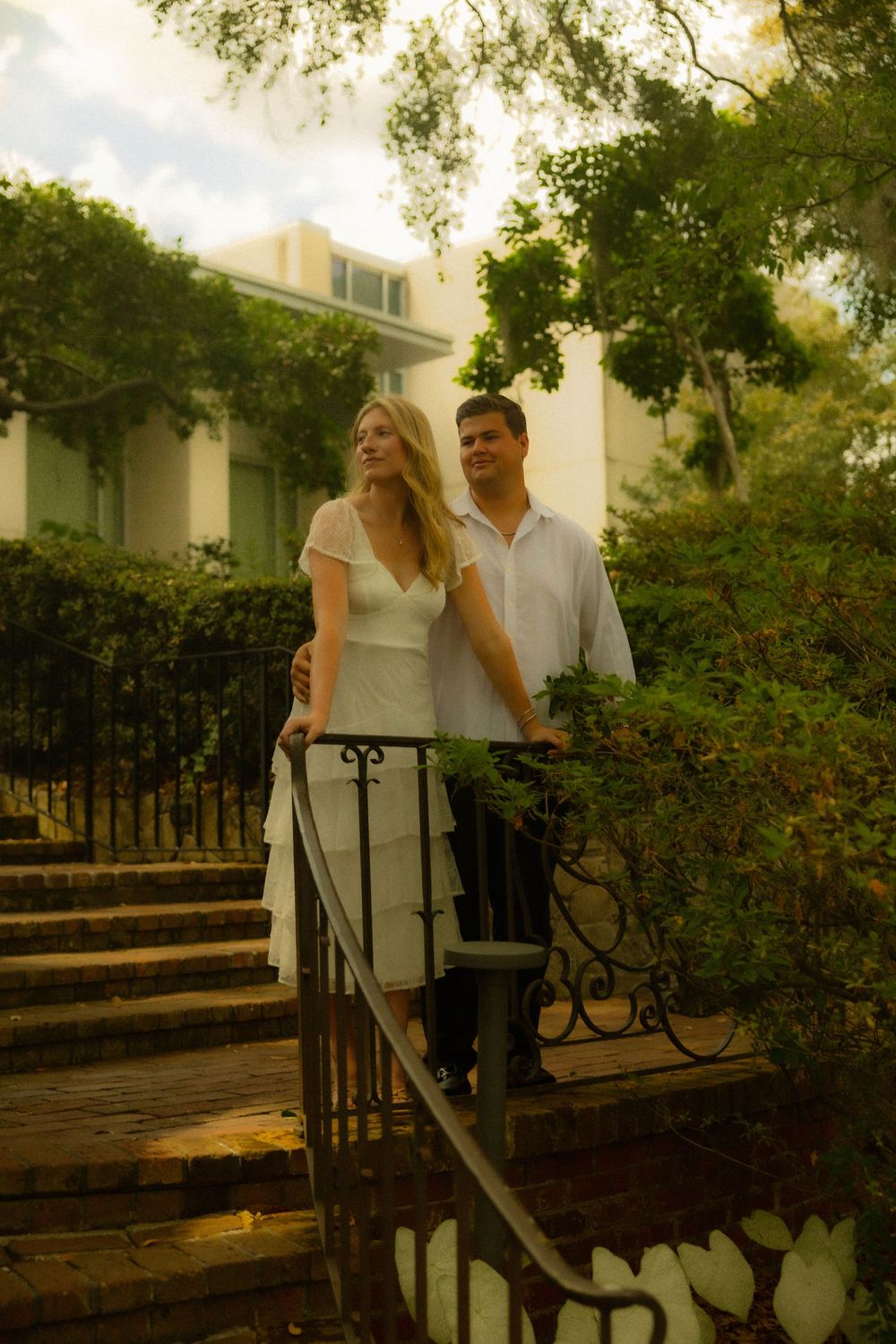 Elegant couple in white attire standing on brick steps by wrought iron railing outside a lush garden estate.