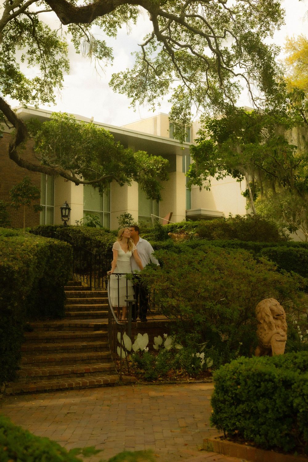 Romantic couple sharing a kiss on garden stairs surrounded by manicured hedges and large oak trees at estate.