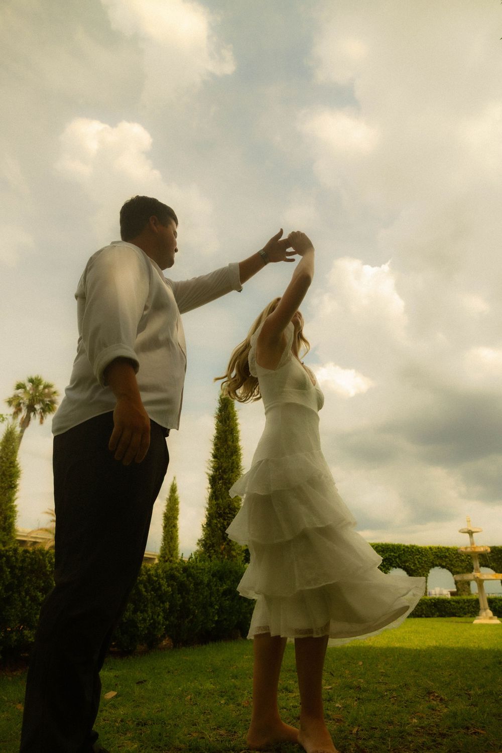 Couple dancing outdoors on green lawn with cypress trees and cloudy sky, woman in white tiered dress being twirled by man.