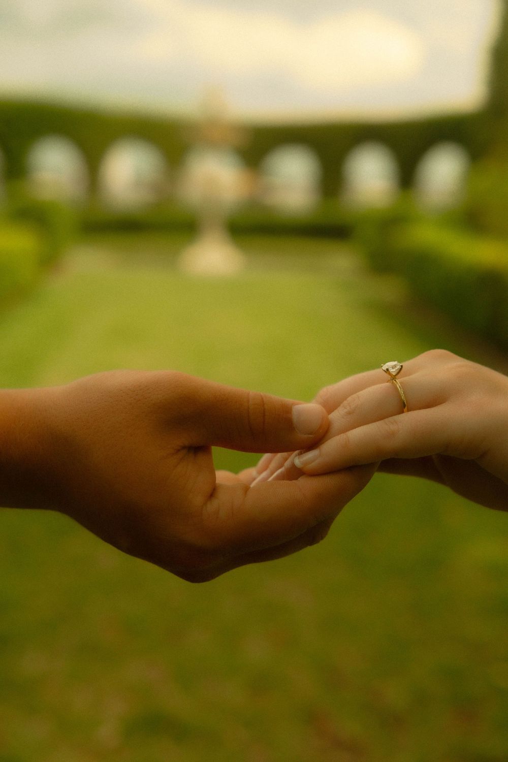 Two hands gently touching, one wearing a gold engagement ring, in a lush green garden with arched architecture.