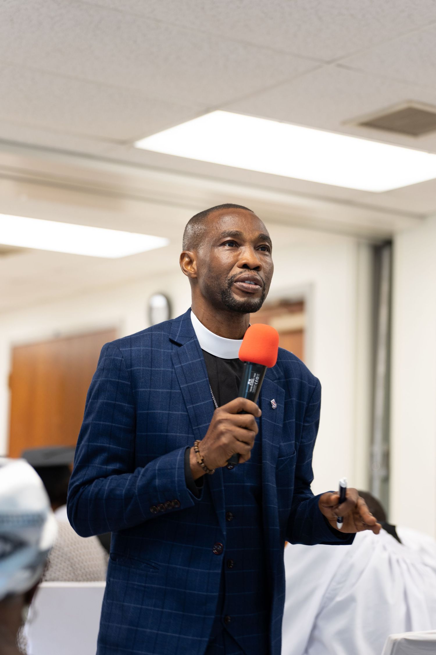 Man in blue plaid suit holding red microphone speaking at indoor event.