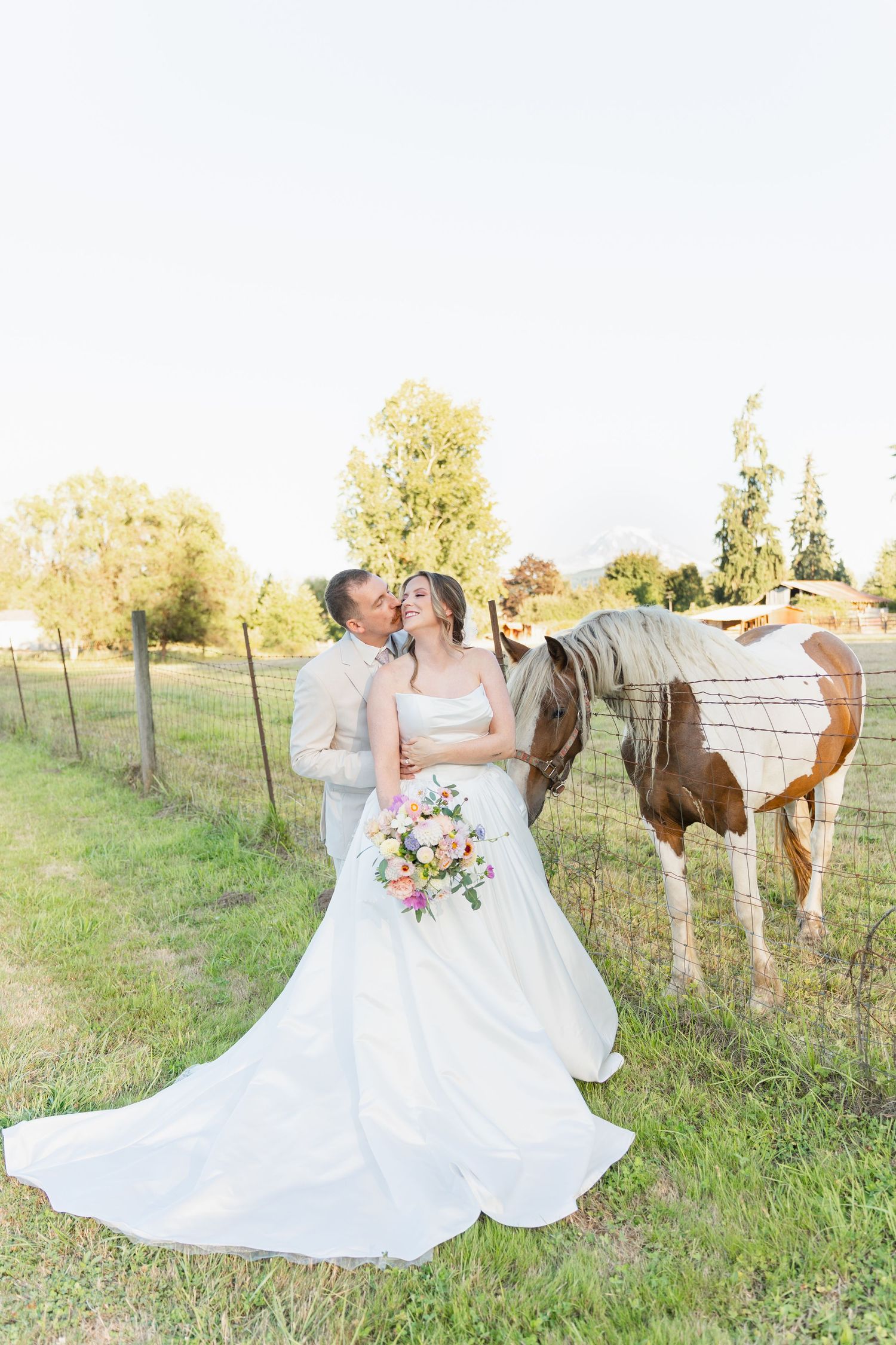 Bride and groom share a kiss beside a paint horse in a sunny green pasture, bride holding a colorful bouquet.