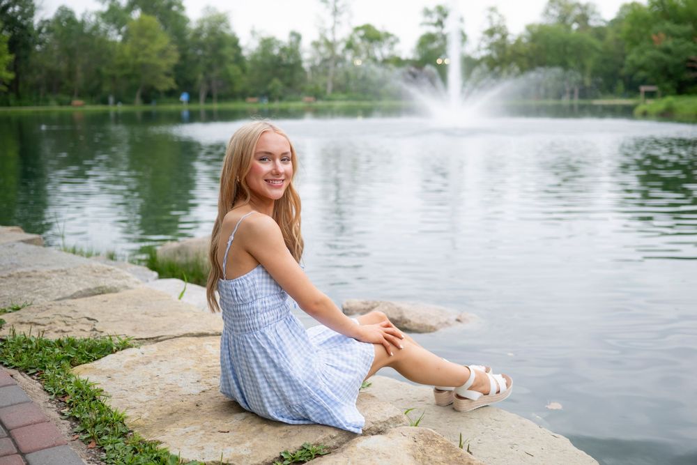 A light blue sundress and white sandals complement the peaceful pond setting with a fountain in the background.