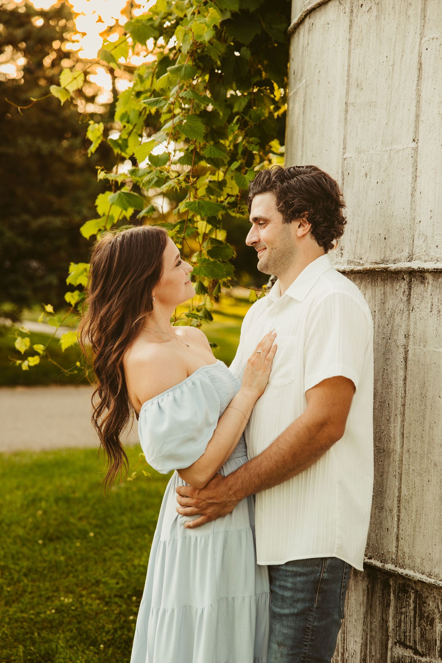 A romantic couple embraces against a silo at sunset with green foliage in the background. Novi Northville Portrait Photographer