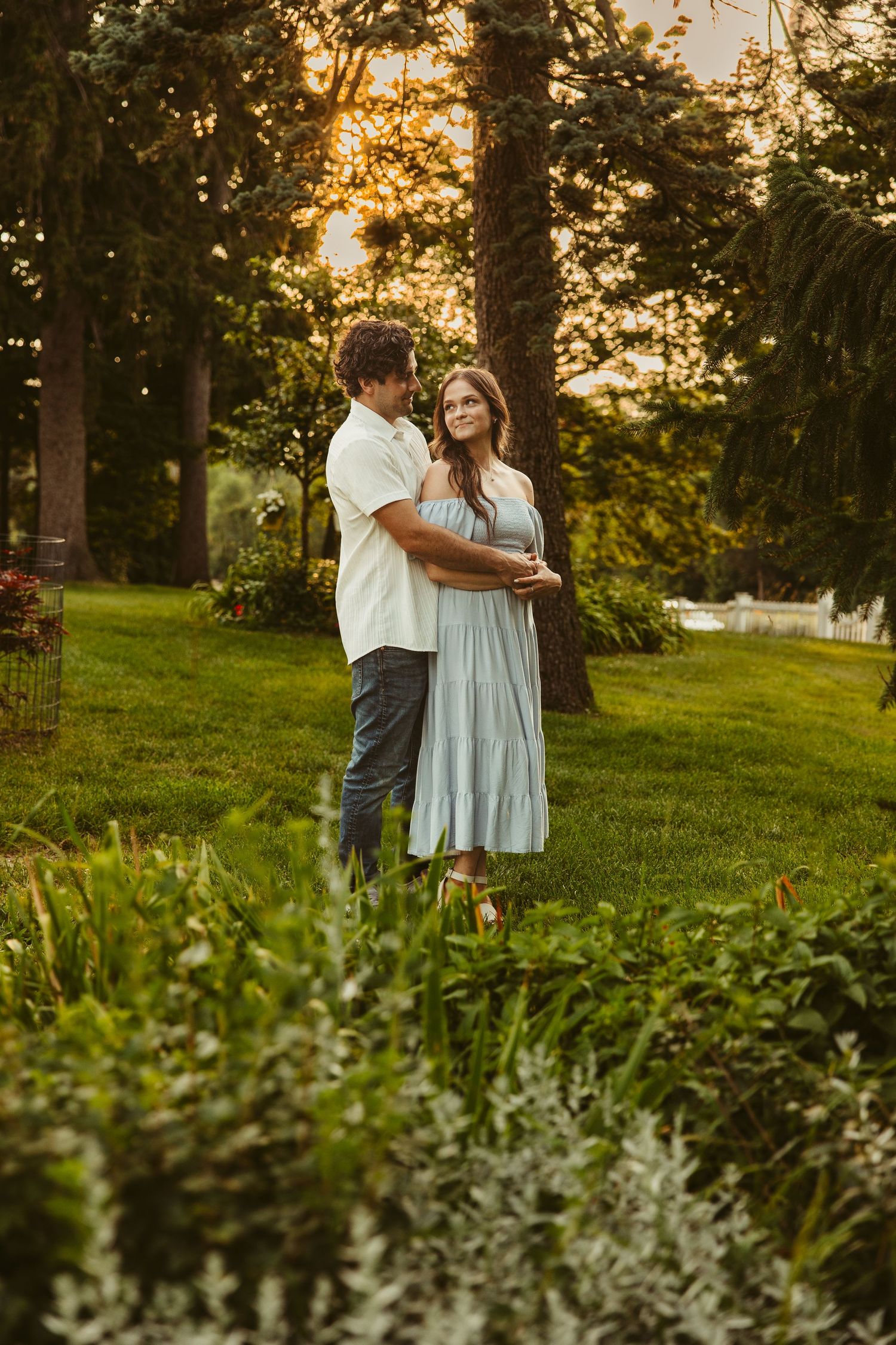 A romantic couple embraces at sunset in a lush garden setting with golden light filtering through tall trees.
