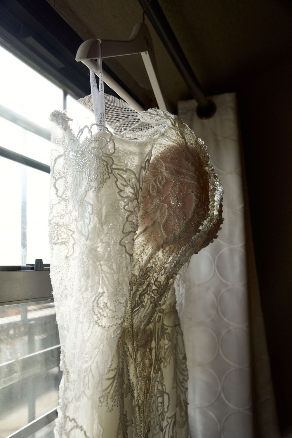 White lace wedding dress hanging by a window with natural light streaming in.
